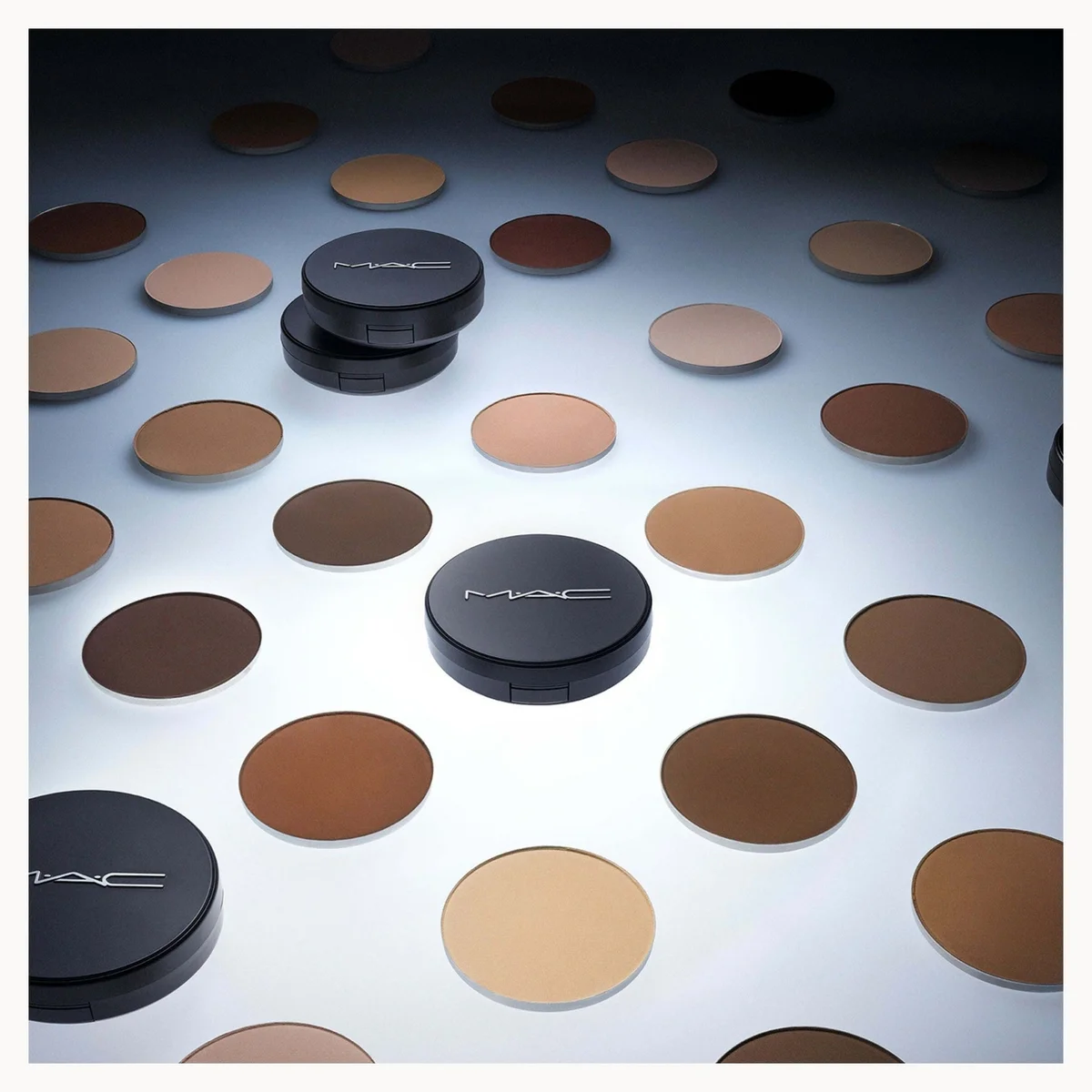 MAC makeup compact powders in various shades of brown, beige, and black, arranged on a reflective white surface with circular pans of matching powder in the background.