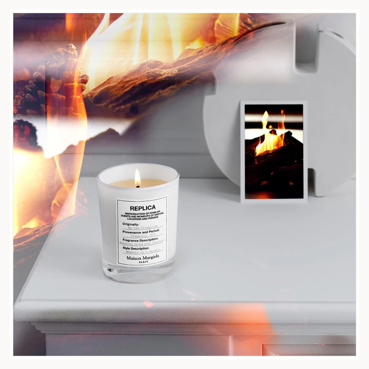 A white glass jar candle with a lit wick sits on a white surface next to a small printed card showing a flame scene. The background is a collage of warm orange and brown flames.