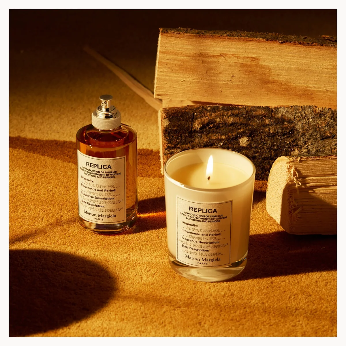A Replica candle and matching perfume bottle styled with wood props on a warm, sandy surface under dramatic golden lighting. The candle is lit, and both products display the 'REPLICA' brand label.