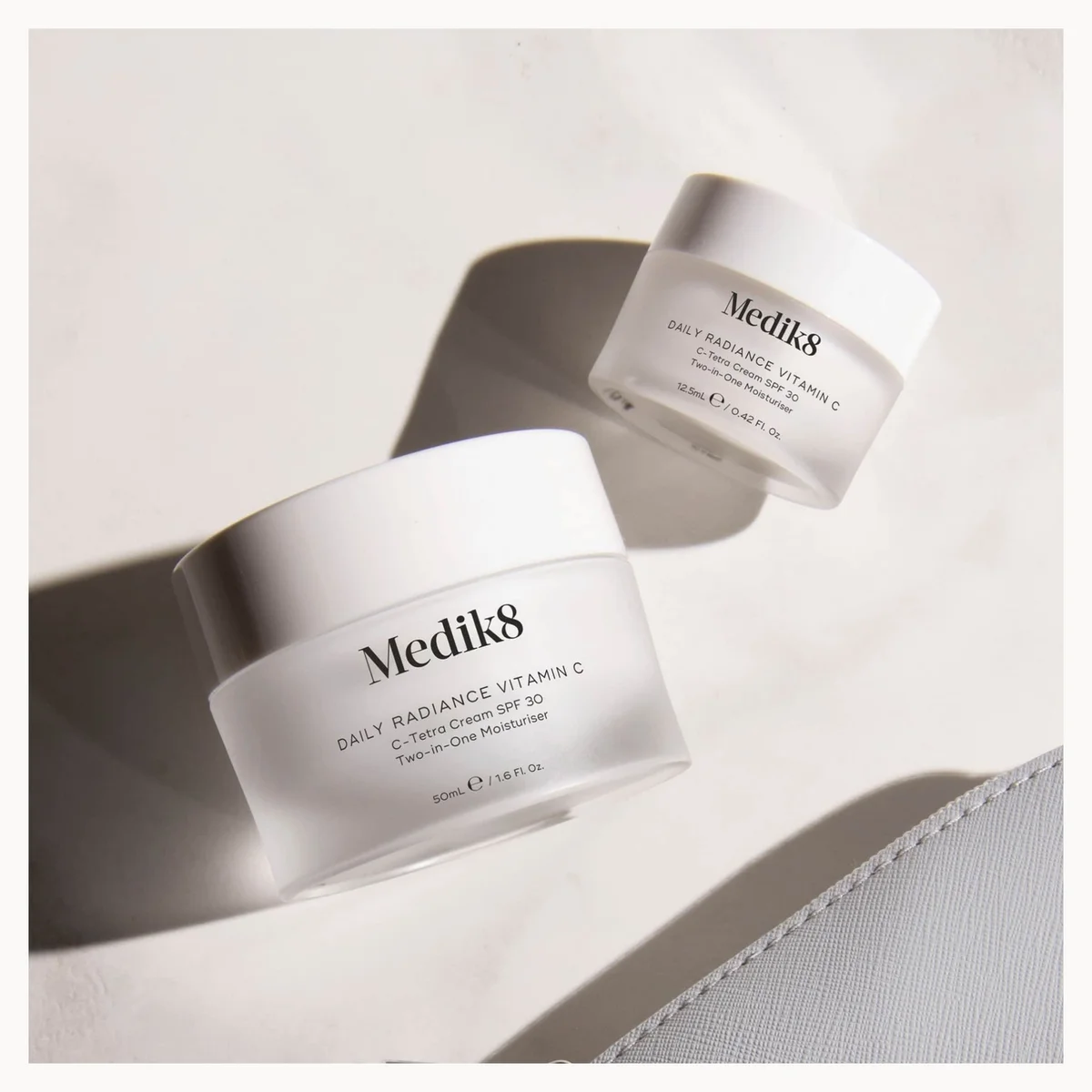Two white Medik8 skincare jars with frosted glass bodies and white lids, photographed on a light neutral surface with strong directional shadows. The front jar is larger and shows the text 'Medik8 Daily Radiance Vitamin C' and 'SPF 30 / PA++++'.
