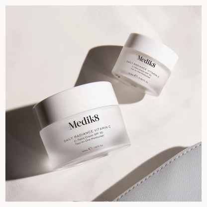Two white Medik8 skincare jars with frosted glass bodies and white lids, photographed on a light neutral surface with strong directional shadows. The front jar is larger and shows the text 'Medik8 Daily Radiance Vitamin C' and 'SPF 30 / PA++++'.