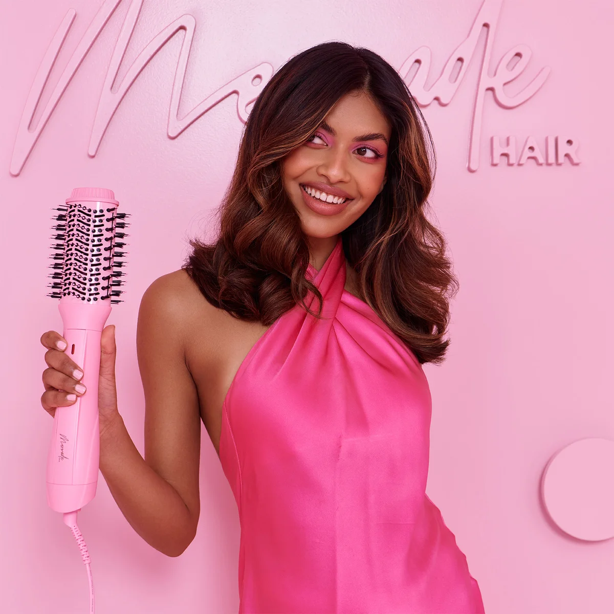 A woman with wavy brown hair holds a pink hair styling brush against a pink background with the words 'Made HAIR' visible on the wall.