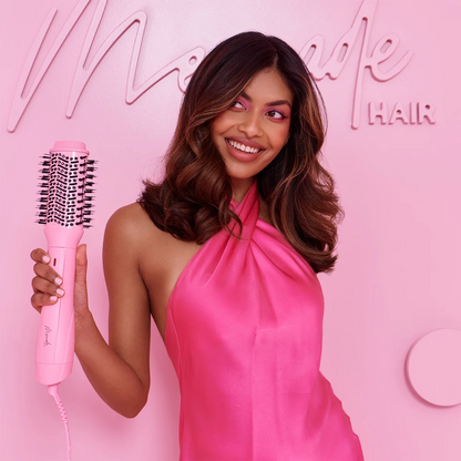 A woman with wavy brown hair holds a pink hair styling brush against a pink background with the words 'Made HAIR' visible on the wall.