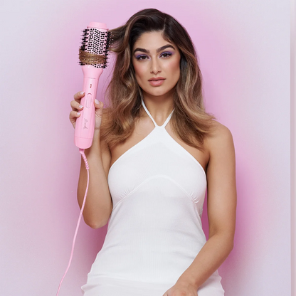 A woman with long wavy brown hair holds a pink round hair brush in her hand, wearing a white halter top against a soft pink background.