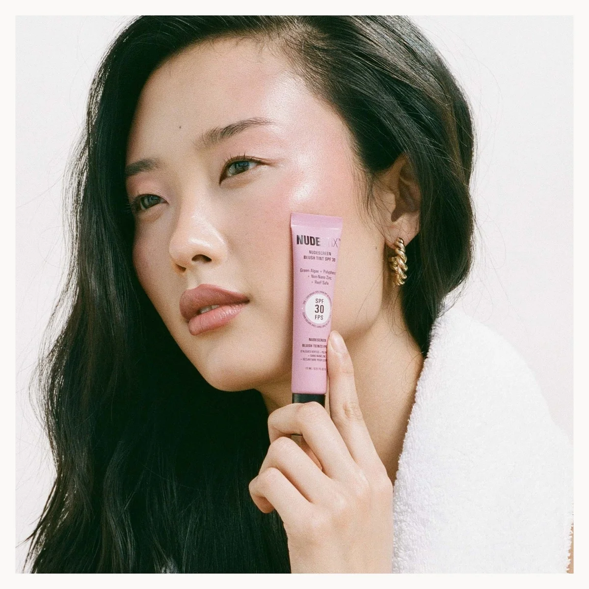 A woman with long dark hair holds a pink tube of AVISOI sunscreen against her cheek, wearing a white robe against a clean white background.