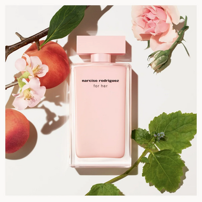 A Narciso Rodriguez 'for her' perfume bottle in a soft pastel pink color, styled in a flat-lay arrangement with fresh peaches, green leaves, and pink roses on a clean white background.