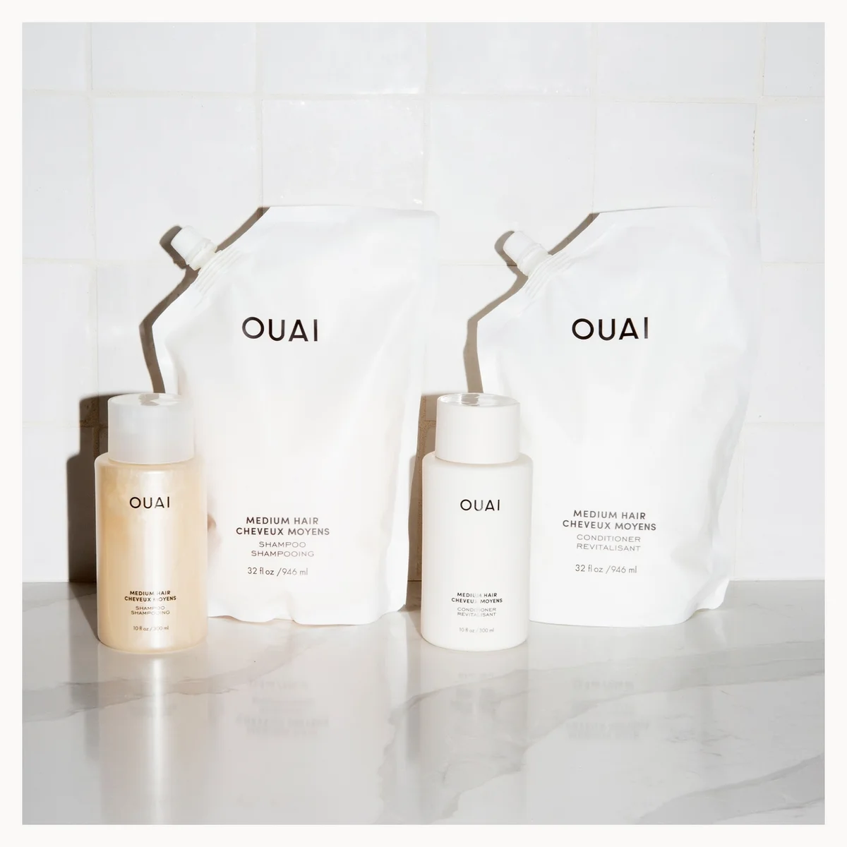 OUAI hair care set featuring two white refill pouches and two white bottles with beige liquid containers, arranged on a light marble surface against a white tiled background.