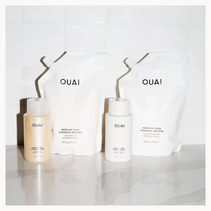 OUAI hair care set featuring two white refill pouches and two white bottles with beige liquid containers, arranged on a light marble surface against a white tiled background.