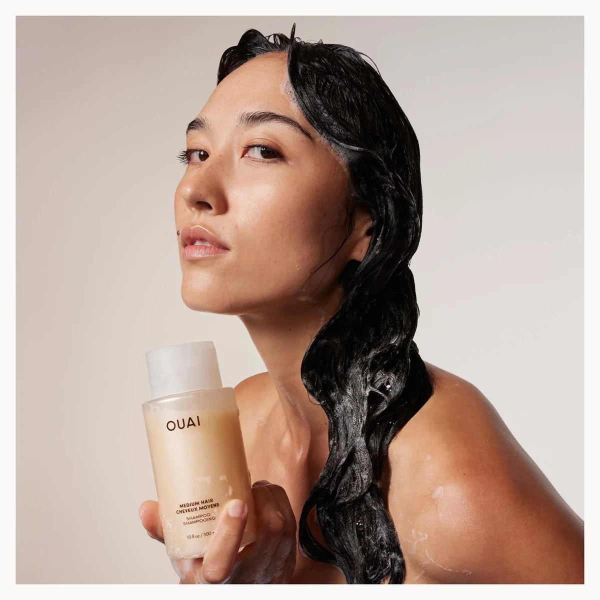 A woman with wet dark hair holds a bottle of OUAI shampoo in a studio setting with a warm beige background.