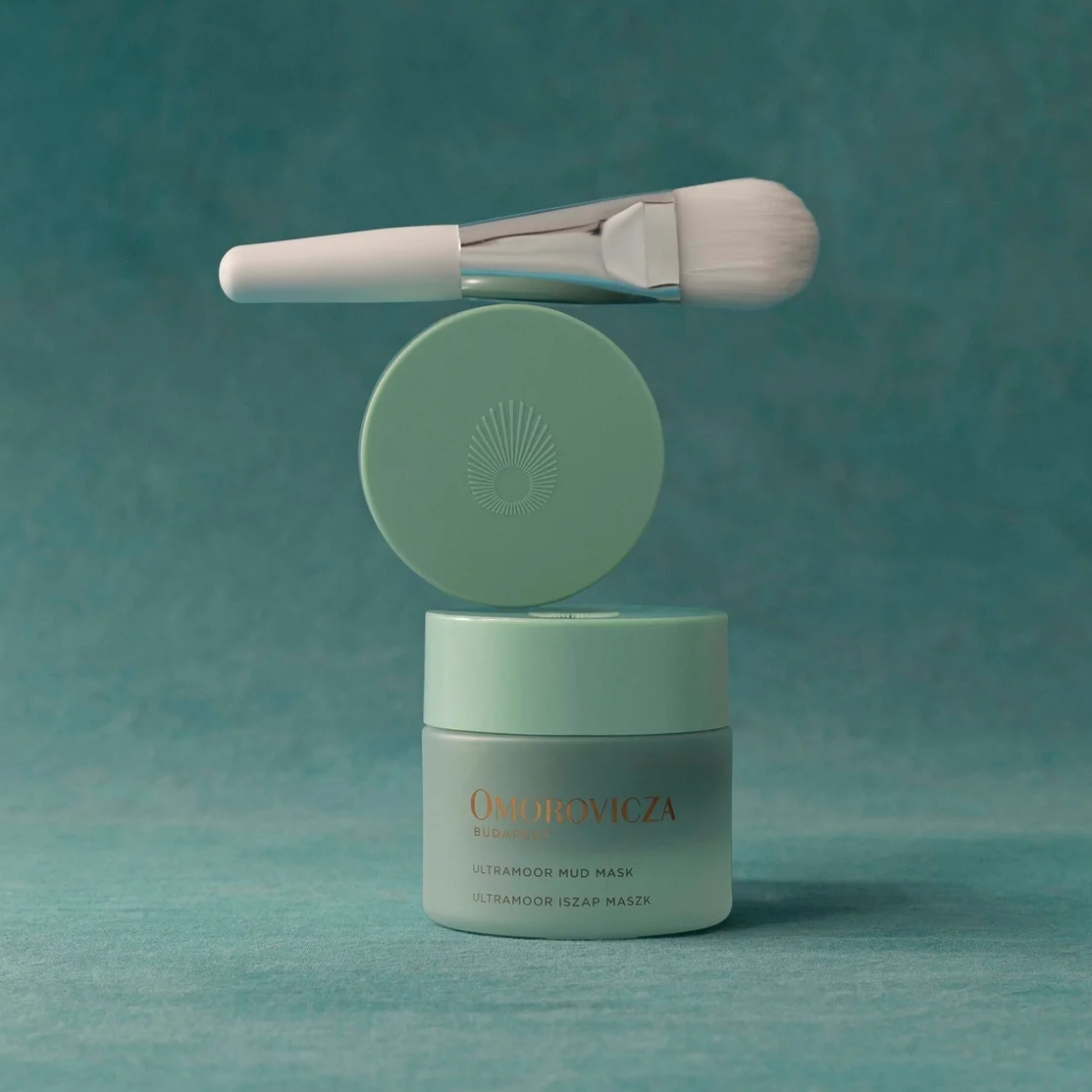 A cosmetic jar labeled 'OCEANVAZA' with a matching round lid leaning behind it and a white applicator brush resting across the top, all set against a teal studio background.