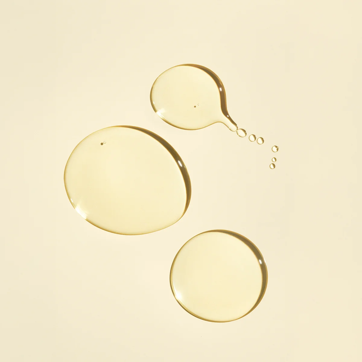 Three translucent golden-yellow liquid droplets on a warm cream background, with one larger droplet splitting into smaller ones.