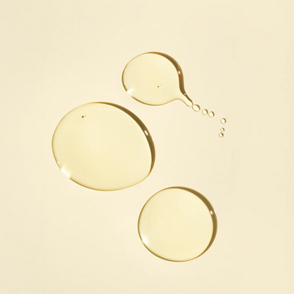 Three translucent golden-yellow liquid droplets on a warm cream background, with one larger droplet splitting into smaller ones.