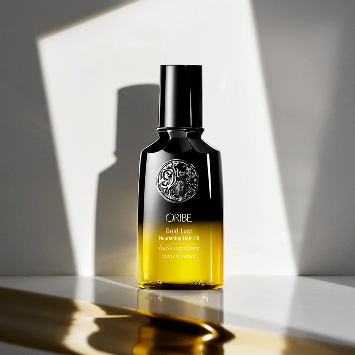 A bottle of Oribe Gold Lust Nourishing Men Oil standing on a reflective surface with dramatic geometric shadows. The bottle is black at the top and transitions to a rich golden yellow at the bottom, with the Oribe logo clearly visible.