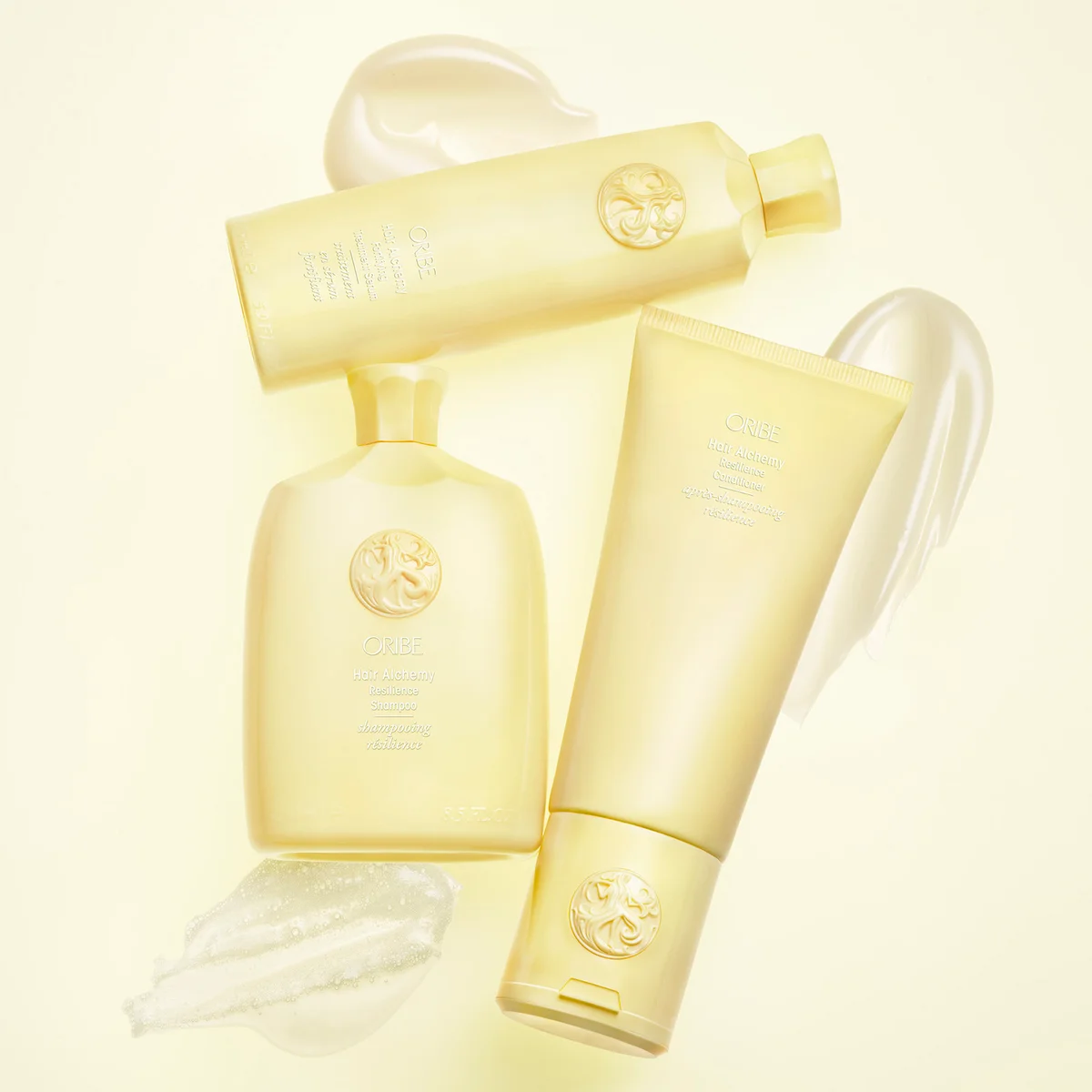 Three pale yellow cosmetic bottles and tubes arranged on a light cream background with translucent gel swatches. The embossed logo and text indicate CeraVe.