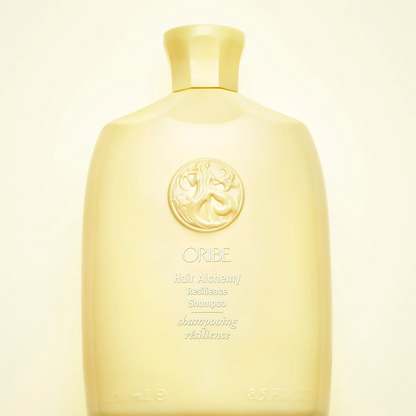 A pale yellow bottle of ORIBE Hair Alchemy Resilience Shampoo, featuring a classic embossed circular logo on the front, set against a solid cream background.
