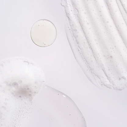 A close-up abstract product shot showing a clear, milky liquid with fine bubbles, including a circular droplet and a smudge of the same product on a very light gray background.
