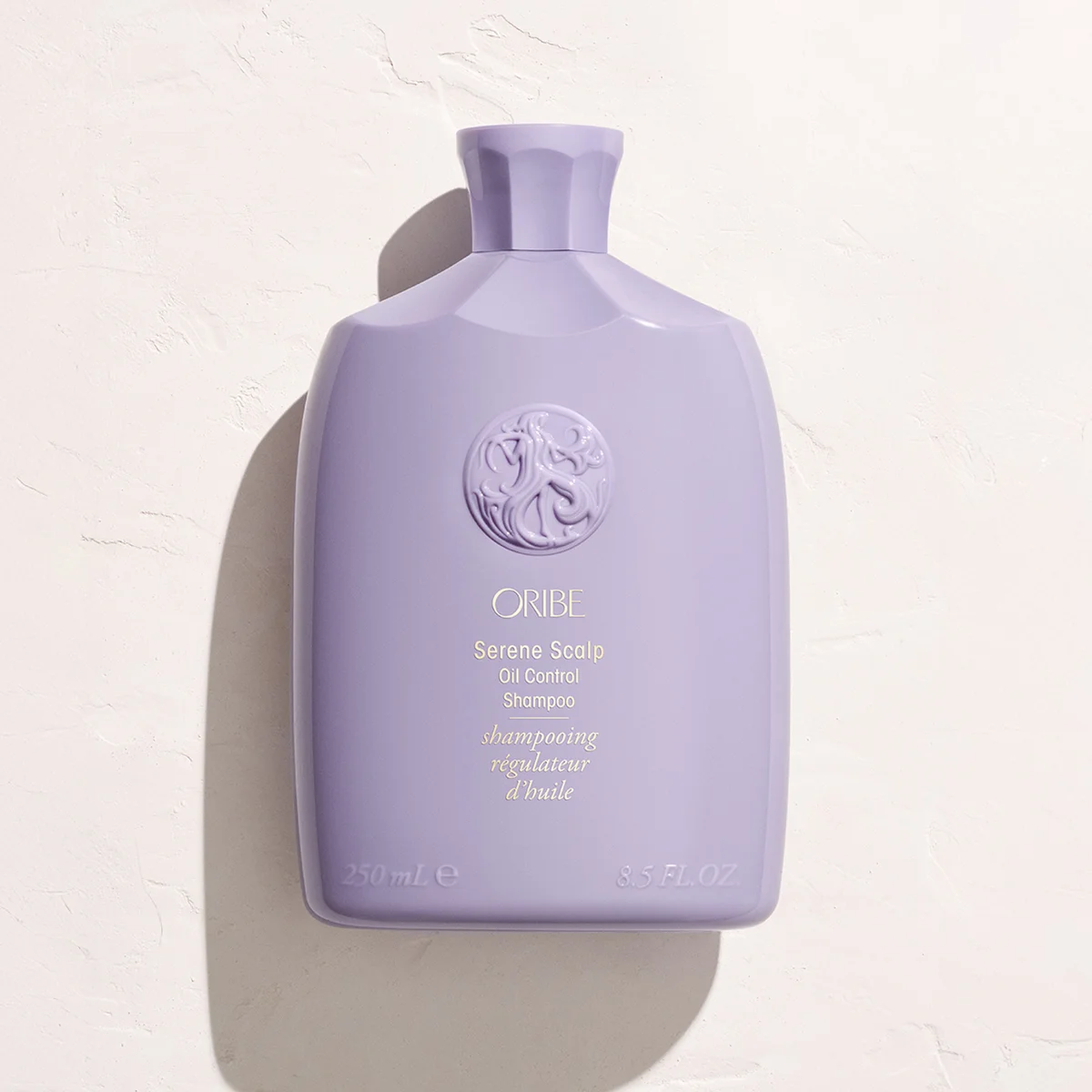 A lavender purple bottle of Oribe Serene Scalp Oil Control Shampoo, photographed from a high angle against a textured off-white background with a soft shadow.