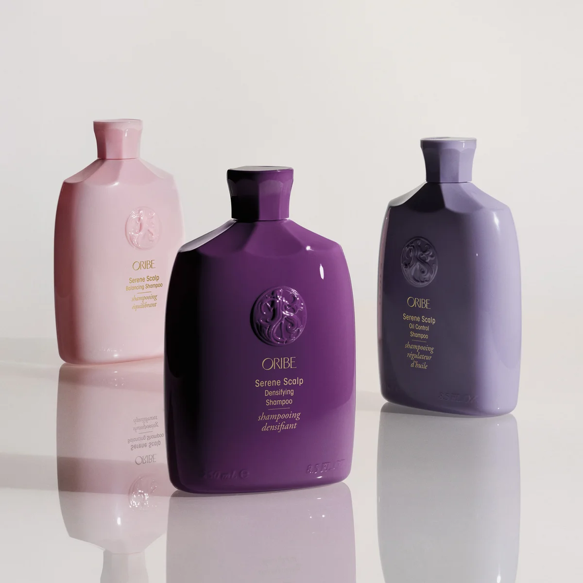 Three Oribe shampoo bottles in soft pink, deep purple, and muted lavender, arranged on a glossy white reflective surface against a light neutral background.