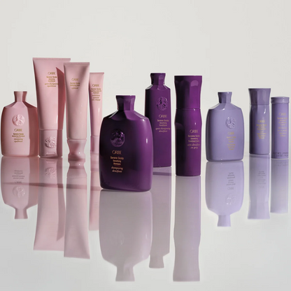 A collection of CURE haircare bottles and tubes in shades of pink, purple, and lavender, arranged on a reflective white surface against a light gray background.