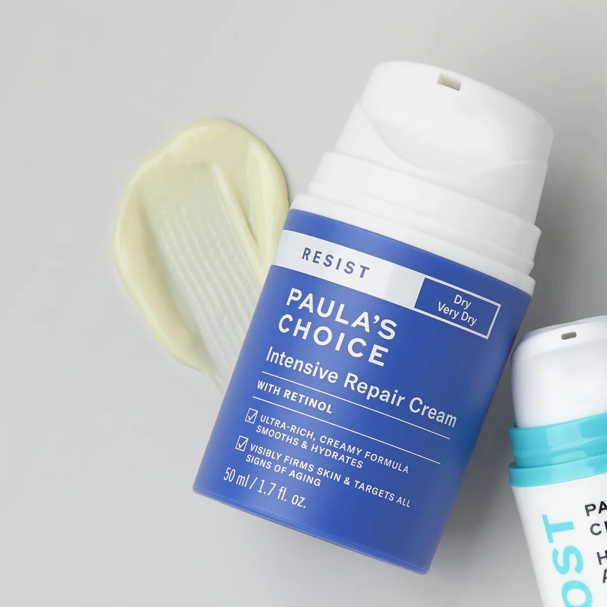 A bottle of Paula's Choice Resist Intensive Repair Cream with Retinol lies on a light gray surface next to a smear of pale yellow cream and part of another skincare bottle.