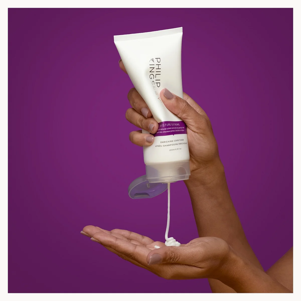Hand holding a tube of Philip B hair product with visible cream on a purple background