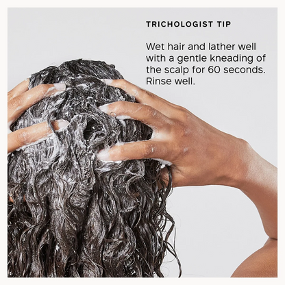 A close-up of a person's hands washing dark curly hair with shampoo lather, with text reading 'TRICHOLOGIST TIP' and instructions on how to wet and knead the hair. The image is presented on a white background with a gray inset panel.