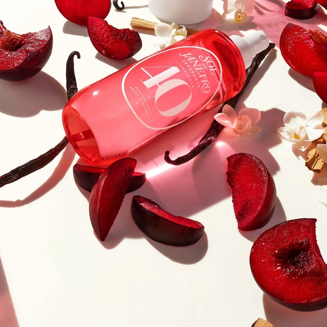 A pink translucent bottle labeled '10 ANTS' and 'PLUM' is arranged on a white surface with sliced red plums, vanilla beans, and small flowers around it.