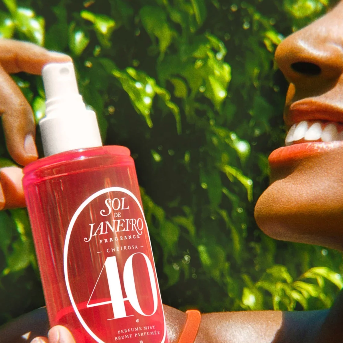 A person outdoors holds a translucent pink bottle labeled 'Sol de Janeiro Perfume Air 40' in front of a bright green leafy background.