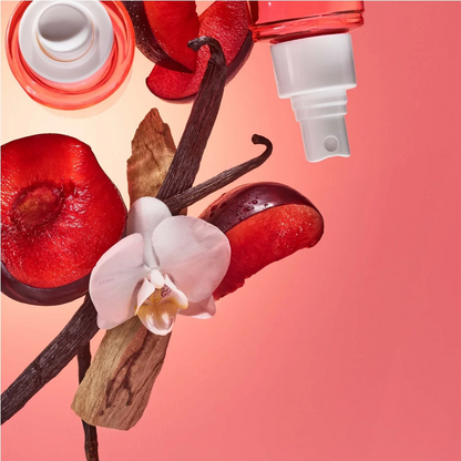 A perfume or fragrance spray bottle with a white atomizer and a red cap, styled with red fruit slices, a vanilla bean, a vanilla pod, and a white orchid flower on a pink background.
