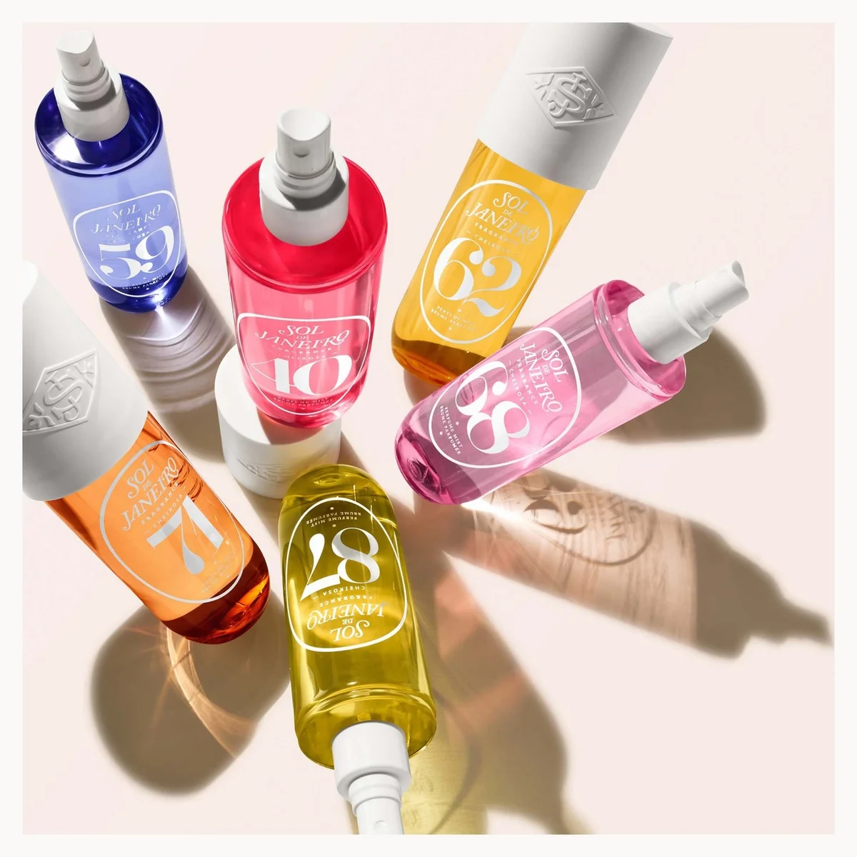 Six cosmetic spray bottles from the brand KIMCHOO, arranged in a circle on a light beige background with soft shadows. The bottles contain colorful liquids in shades of blue, red, yellow, and pink, with white caps and white labels showing numbers 39, 10, 62, 17, 28, and 33.