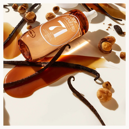 A bottle of Sol-de-Janeiro-Cheirosa hair and body mist with a white dropper cap, styled diagonally on a white background with vanilla beans, hazelnuts, and a drizzle of amber liquid.