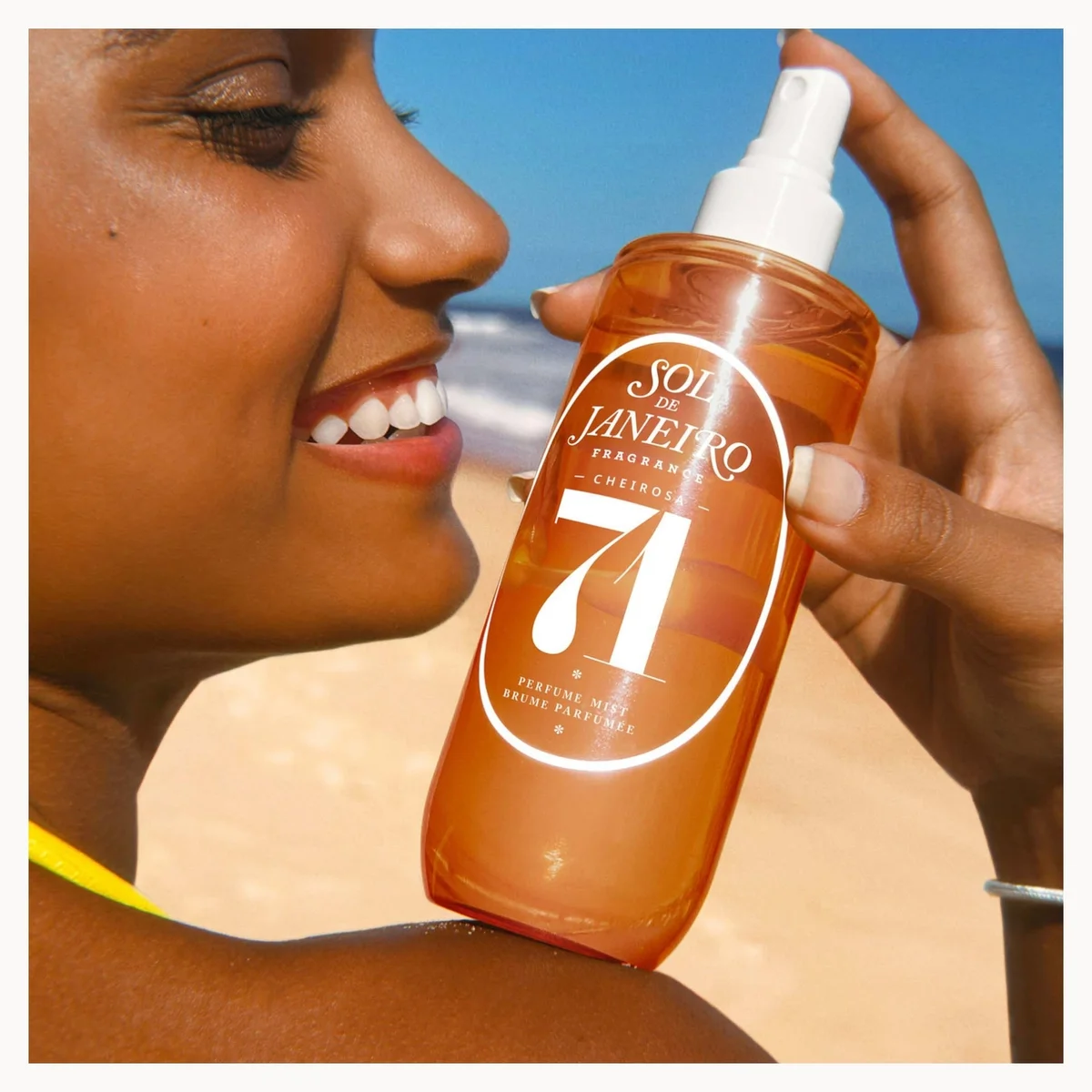 A woman at the beach holds a bottle of Sol de Janeiro Natural 71 Sunscreen Spray. The bottle is a translucent amber orange with a white pump and a large white oval logo on the front.