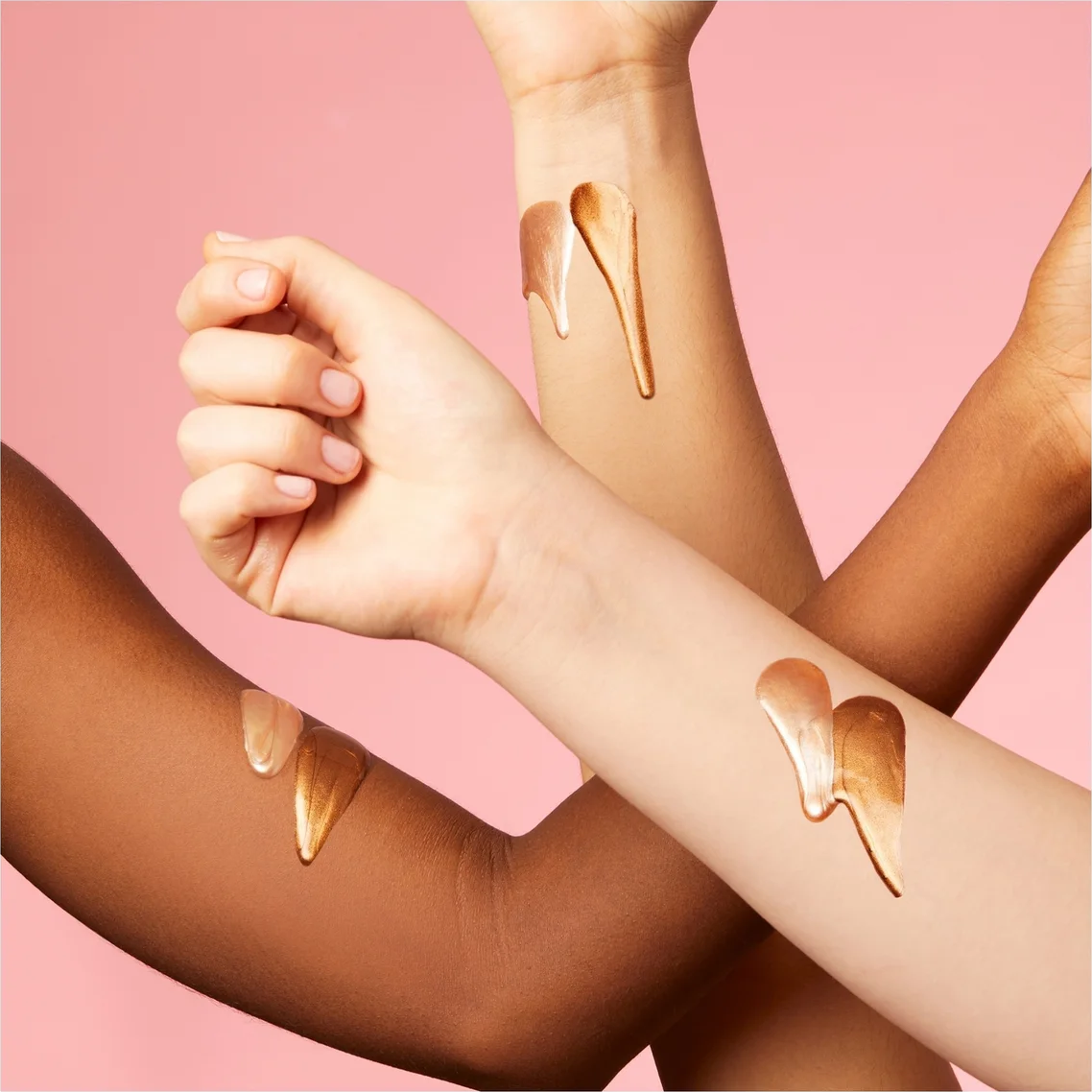 Three crossed human arms against a soft pink background, each showing a glossy, metallic gold liquid smear on the skin.