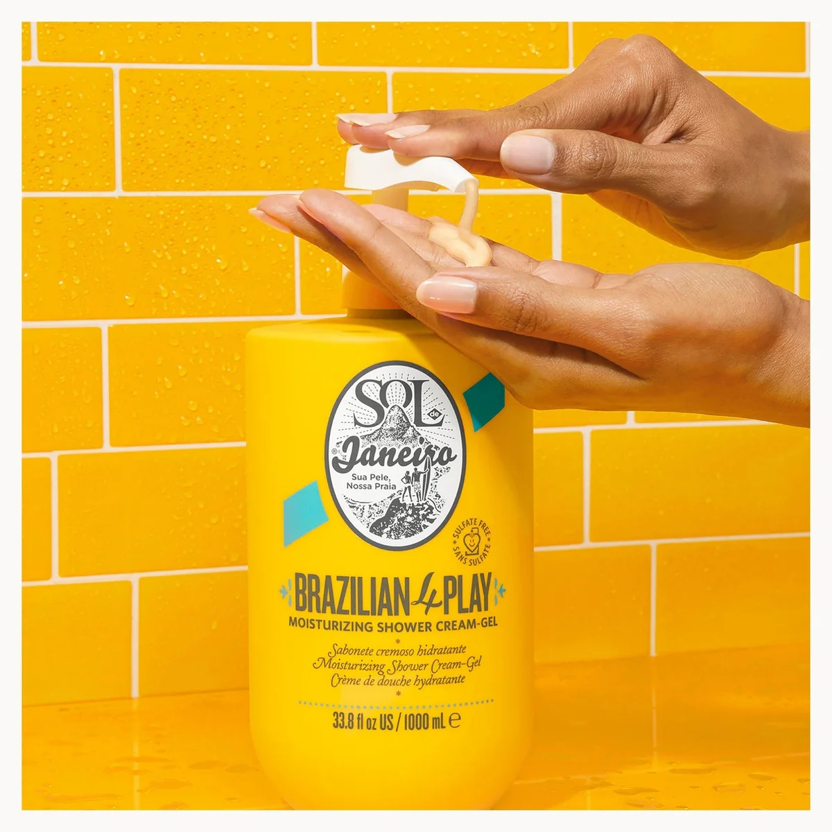 A person's hands are shown dispensing cream from a yellow bottle of Sol Janeiro Brazilian 4-Play moisturizing shower cream gel against a yellow tiled background.
