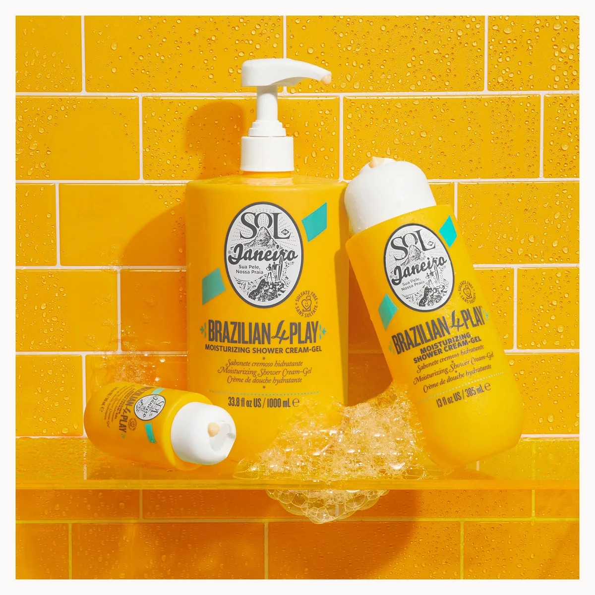 Three yellow sol de janeiro Brazilian-4-Play hair care bottles arranged on a clear shelf against a yellow tiled background with water droplets and foam.