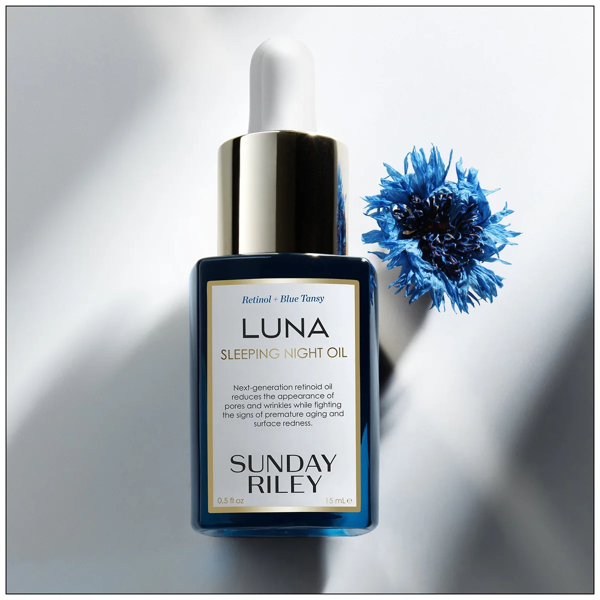 Sunday Riley Luna Sleeping Night Oil in a dark blue glass dropper bottle with a gold cap, styled next to a blue cornflower on a white background with soft shadows.