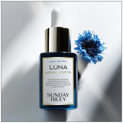 Sunday Riley Luna Sleeping Night Oil in a dark blue glass dropper bottle with a gold cap, styled next to a blue cornflower on a white background with soft shadows.