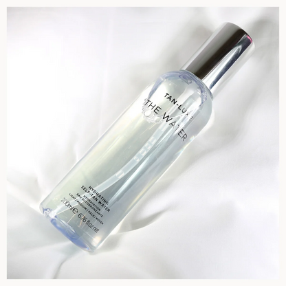 A clear cylindrical bottle of TAI LUXE THE WATER makeup setting spray with a reflective silver cap, resting on a white fabric background.
