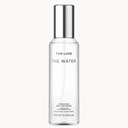 A clear cylindrical bottle of Tan-Luxe 'The Water' hydrating self-tan water with a reflective silver cap, centered on a light gray background.