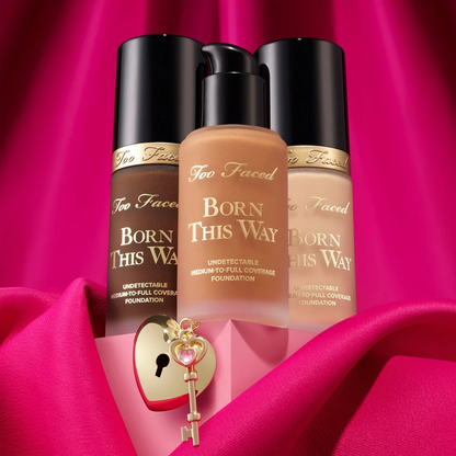 Three Too Faced Born This Way foundation bottles arranged together on draped hot pink fabric, with a small gold key and pink box in the foreground.