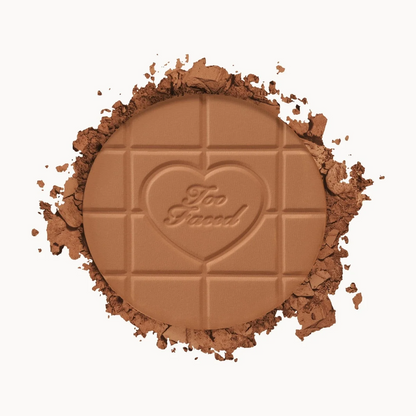 Too Faced chocolate bar shaped pressed powder makeup on a clean white background, surrounded by crushed matching brown powder.