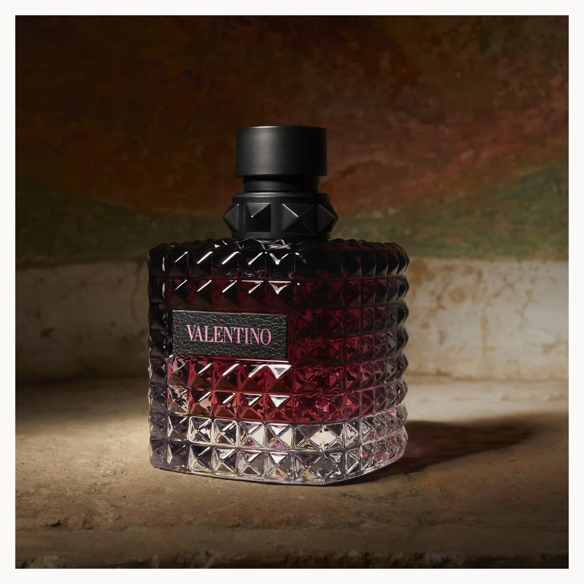 A textured glass perfume bottle with a faceted diamond pattern, filled with deep burgundy liquid and featuring a black cap. The brand name 'VALENTINO' is printed on a black label on the front.