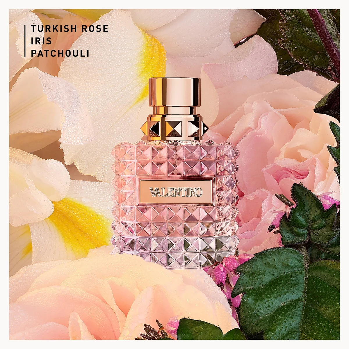 A Valentino perfume bottle labeled 'Turkish Rose Iris Patchouli' placed among soft pink roses and green leaves. The bottle is faceted pink glass with a metallic rose-gold cap and a gold-toned front plaque.