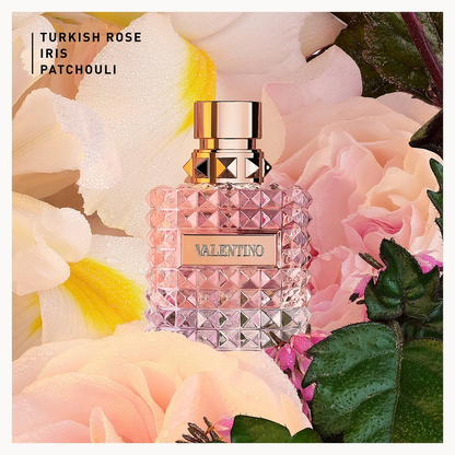 A Valentino perfume bottle labeled 'Turkish Rose Iris Patchouli' placed among soft pink roses and green leaves. The bottle is faceted pink glass with a metallic rose-gold cap and a gold-toned front plaque.