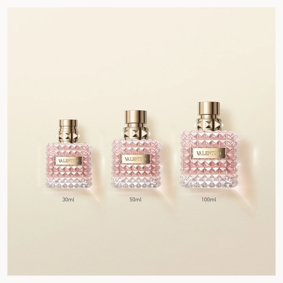 Three Valeria Rossi perfume bottles in 30ml, 50ml, and 100ml sizes lined up on a light beige background. The bottles are clear with a textured pink and white beaded pattern, topped with gold caps, and feature a gold brand label.