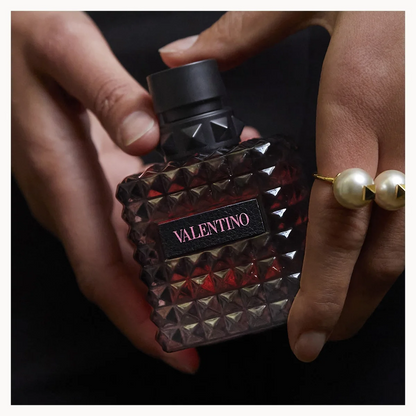 A close-up of a person holding a Valentino perfume bottle with both hands. The bottle is dark brown with a textured, faceted pyramid pattern and a black cap.