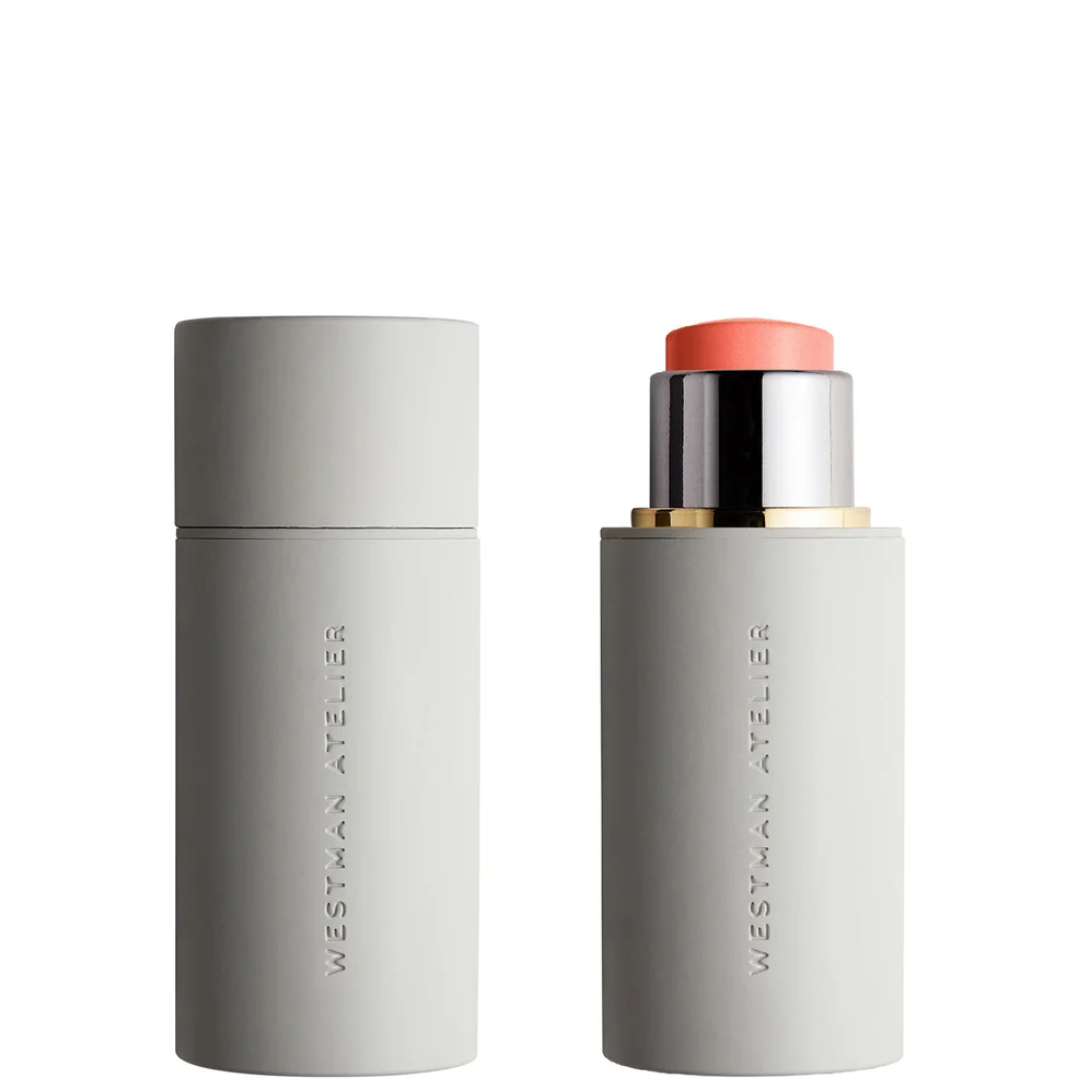 Westman Atelier cosmetic stick product shown closed in a matte light gray cylinder with the cap on, and open with the coral-pink product exposed, a metallic silver cap, and a small gold ring, on a white background.
