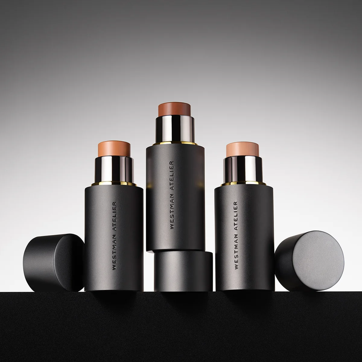 Three cosmetic sticks with visible 'Westman Atelier' branding on a gradient background