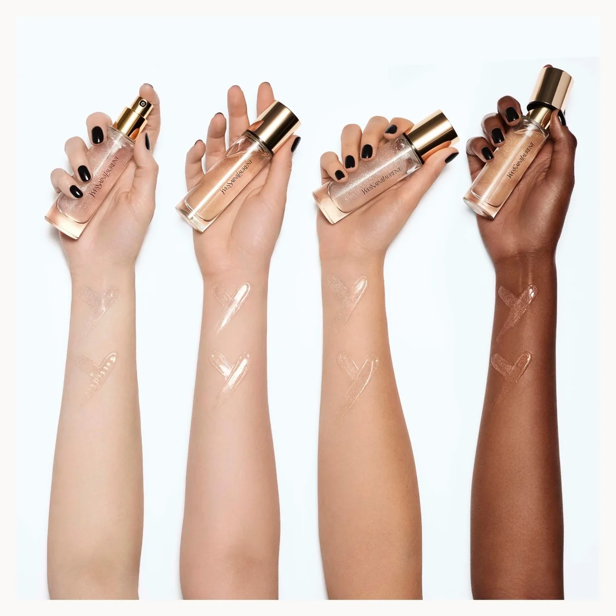Four hands of different skin tones holding clear glass bottles with gold caps, each swatched with a transparent liquid on their forearm against a white background.