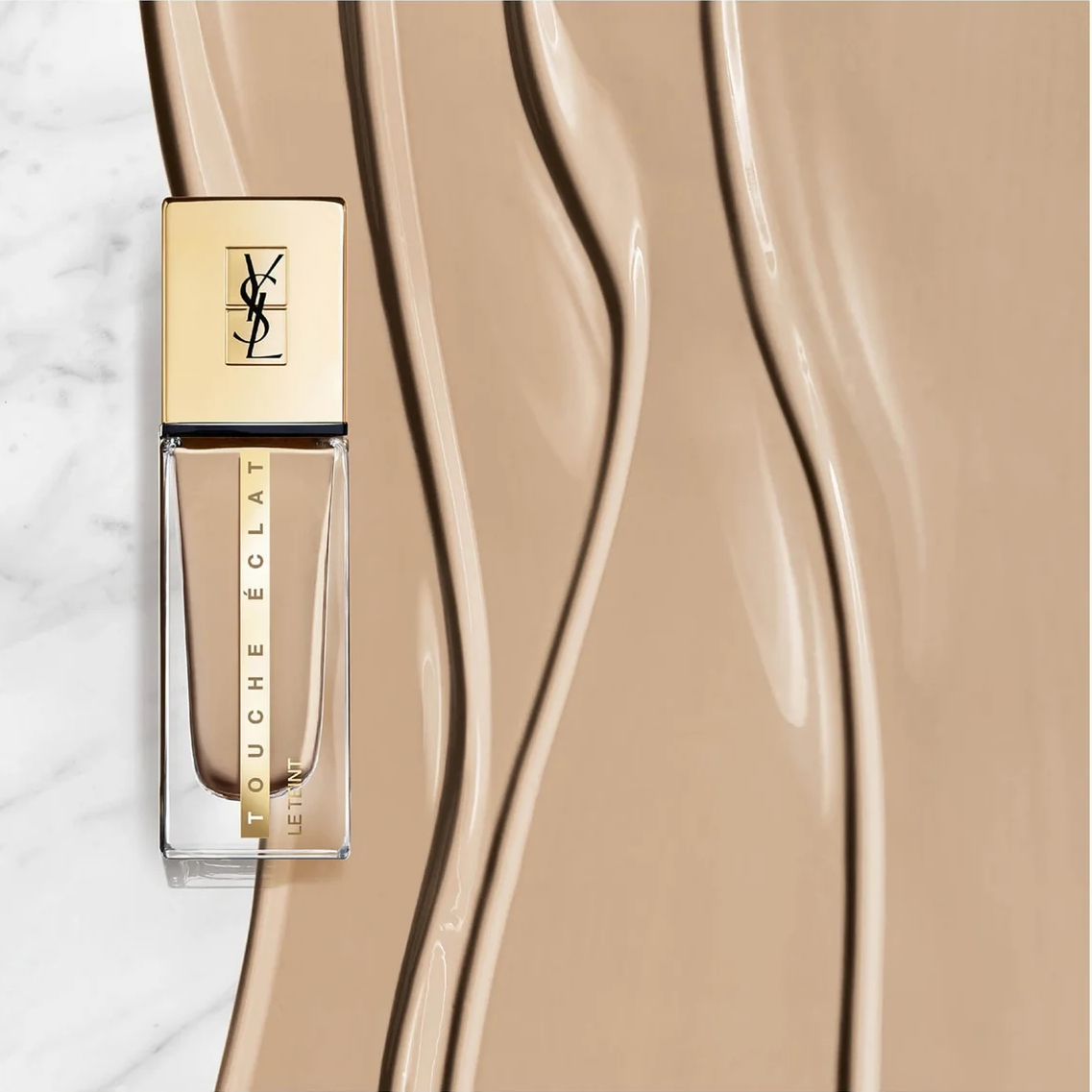 YSL Touche Eclat liquid foundation bottle in a warm beige shade, shown next to a large, glossy smear of the matching foundation on a light marble background.